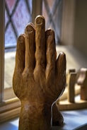 A wooden sculpture of a hand, carefully carved with detailed features, is positioned upright against a textured window. The background reveals blurred shapes of additional wooden items and a crosshatched window pattern.