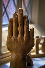 A wooden sculpture of a hand, carefully carved with detailed features, is positioned upright against a textured window. The background reveals blurred shapes of additional wooden items and a crosshatched window pattern.