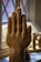 A wooden sculpture of a hand, carefully carved with detailed features, is positioned upright against a textured window. The background reveals blurred shapes of additional wooden items and a crosshatched window pattern.