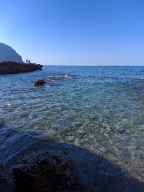 Clear blue ocean water extends toward the horizon under a bright sky. A rocky shoreline is visible on the left with a few people standing on the rocks, enjoying the view. More people are seen in the water further out, possibly engaging in water activities.