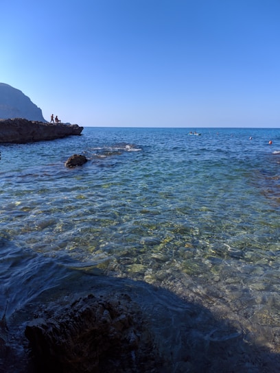 Clear blue ocean water extends toward the horizon under a bright sky. A rocky shoreline is visible on the left with a few people standing on the rocks, enjoying the view. More people are seen in the water further out, possibly engaging in water activities.