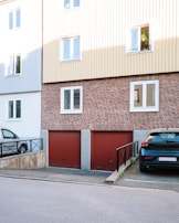 A residential building with a brick facade and beige siding on the upper floors. The ground level features two red garage doors. Parked cars are visible, one in a parking space beside a ramp with black railings. Windows on the upper floor reflect daylight, with one open window displaying hanging plants.