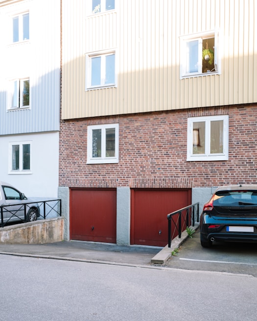 A residential building with a brick facade and beige siding on the upper floors. The ground level features two red garage doors. Parked cars are visible, one in a parking space beside a ramp with black railings. Windows on the upper floor reflect daylight, with one open window displaying hanging plants.