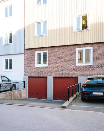 A residential building with a brick facade and beige siding on the upper floors. The ground level features two red garage doors. Parked cars are visible, one in a parking space beside a ramp with black railings. Windows on the upper floor reflect daylight, with one open window displaying hanging plants.