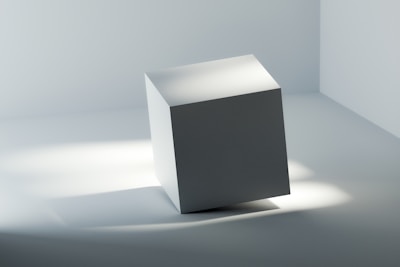 A minimalist scene featuring a single, slightly tilted white cube placed on a smooth surface in a softly lit environment. The lighting creates gentle shadows and gradients on the cube's faces.