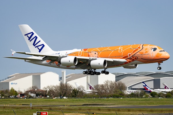A large passenger airplane with distinctive orange and white artwork on its fuselage is flying above an airfield. The plane belongs to ANA, as indicated by the logo on its tail. In the background, there are airport buildings and hangars, with some other aircraft visible. The sky is clear and blue, indicating pleasant weather conditions.