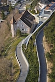 An aerial view of a residential area featuring a large house with a sloping roof surrounded by greenery. The winding pathways lined with shrubs and trees lead up to the house, creating a sense of harmony with the landscape. Several modern buildings are visible, creating a contrast with the traditional architecture of the house.