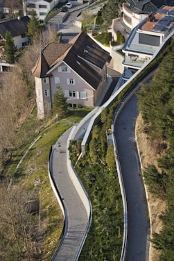 An aerial view of a residential area featuring a large house with a sloping roof surrounded by greenery. The winding pathways lined with shrubs and trees lead up to the house, creating a sense of harmony with the landscape. Several modern buildings are visible, creating a contrast with the traditional architecture of the house.