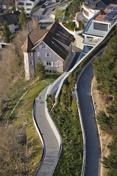 An aerial view of a residential area featuring a large house with a sloping roof surrounded by greenery. The winding pathways lined with shrubs and trees lead up to the house, creating a sense of harmony with the landscape. Several modern buildings are visible, creating a contrast with the traditional architecture of the house.