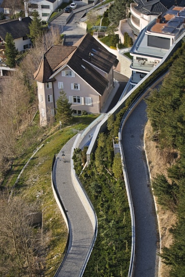 An aerial view of a residential area featuring a large house with a sloping roof surrounded by greenery. The winding pathways lined with shrubs and trees lead up to the house, creating a sense of harmony with the landscape. Several modern buildings are visible, creating a contrast with the traditional architecture of the house.