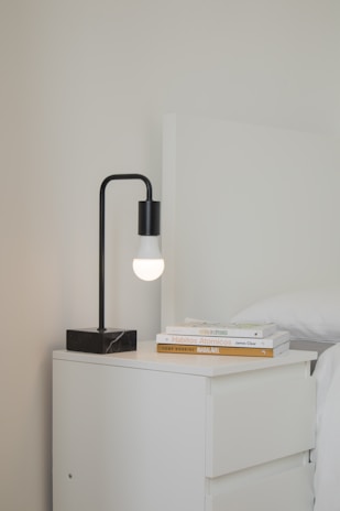 A sleek nightstand with drawers beside a bed, holding a small lamp and a stack of books.