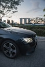 A luxury car with a prominent emblem is parked on the side of a road. In the background, an iconic architectural landmark stands tall against a backdrop of a sunset or early evening sky. Trees and foliage surround the area, creating a serene urban setting.