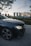 A luxury car with a prominent emblem is parked on the side of a road. In the background, an iconic architectural landmark stands tall against a backdrop of a sunset or early evening sky. Trees and foliage surround the area, creating a serene urban setting.