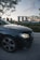 A luxury car with a prominent emblem is parked on the side of a road. In the background, an iconic architectural landmark stands tall against a backdrop of a sunset or early evening sky. Trees and foliage surround the area, creating a serene urban setting.