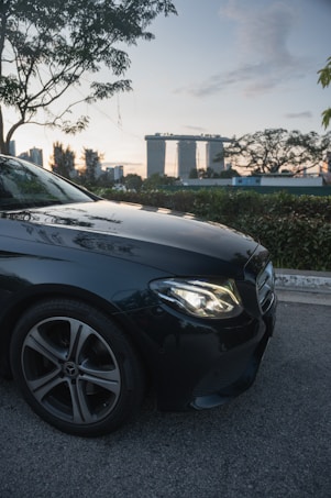 A luxury car with a prominent emblem is parked on the side of a road. In the background, an iconic architectural landmark stands tall against a backdrop of a sunset or early evening sky. Trees and foliage surround the area, creating a serene urban setting.