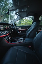The interior of a luxury car features modern black leather seats and a sleek dashboard with illuminated controls. The ambient lighting along the dashboard adds an elegant touch, while the outside view through the windows reveals a green, leafy landscape.