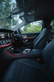 The interior of a luxury car features modern black leather seats and a sleek dashboard with illuminated controls. The ambient lighting along the dashboard adds an elegant touch, while the outside view through the windows reveals a green, leafy landscape.