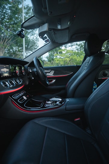 The interior of a luxury car features modern black leather seats and a sleek dashboard with illuminated controls. The ambient lighting along the dashboard adds an elegant touch, while the outside view through the windows reveals a green, leafy landscape.