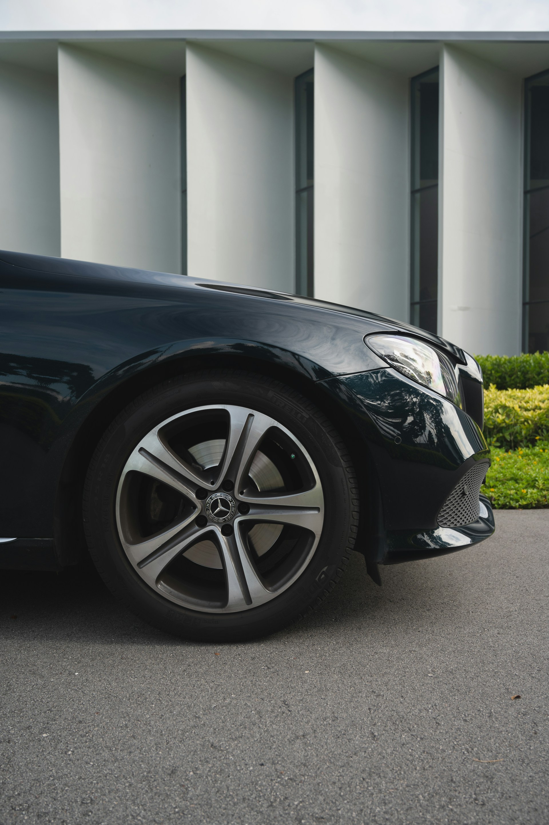 A sleek luxury car is parked on a smooth pavement. The image shows a close-up of the vehicle's front side, featuring its shiny alloy wheel and a distinctive front grille. Tall, modern architectural structures with a symmetrical design are in the background, adding a sophisticated urban atmosphere.