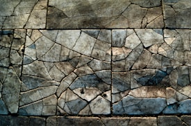 Cracked concrete surface with intricate patterns forming a mosaic-like appearance. The texture resembles a patchwork of jagged tiles with varying shades of gray, brown, and hints of blue, creating an abstract look.