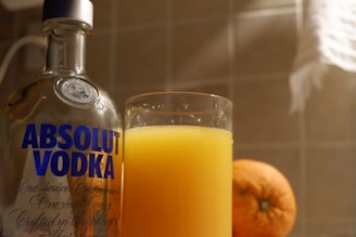 A bottle of Absolut Vodka is positioned next to a glass filled with orange juice, with an orange fruit nearby. The setting appears to be indoors on a tiled surface, illuminated by warm light.