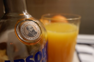 A close-up shot of a bottle of Absolut vodka with its label partially visible, accompanied by a glass filled with orange juice. The focus is on the bottle cap which features an illustration and text indicating Swedish vodka.