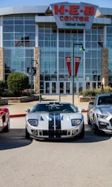 A modern sports car with a sleek design, featuring silver and black stripes, is parked in front of a large building with a sign that says 'H-E-B Center at Cedar Park'. Flanking it are two other cars, and the area appears to be a parking lot. The building has a glass facade with stone accents and banners displaying logos near the entrance.