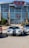 A modern sports car with a sleek design, featuring silver and black stripes, is parked in front of a large building with a sign that says 'H-E-B Center at Cedar Park'. Flanking it are two other cars, and the area appears to be a parking lot. The building has a glass facade with stone accents and banners displaying logos near the entrance.