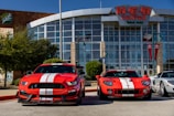 Two bright red sports cars with white racing stripes are parked in front of a large building with glass windows and a sign reading 'H-E-B Center at Cedar Park'. The sports cars contrast sharply against the clear blue sky and the modern architectural design of the building. There are trees and flagpoles in the background, adding a dynamic urban setting to the scene.