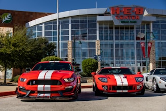 Two bright red sports cars with white racing stripes are parked in front of a large building with glass windows and a sign reading 'H-E-B Center at Cedar Park'. The sports cars contrast sharply against the clear blue sky and the modern architectural design of the building. There are trees and flagpoles in the background, adding a dynamic urban setting to the scene.
