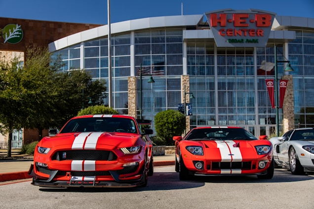 Two bright red sports cars with white racing stripes are parked in front of a large building with glass windows and a sign reading 'H-E-B Center at Cedar Park'. The sports cars contrast sharply against the clear blue sky and the modern architectural design of the building. There are trees and flagpoles in the background, adding a dynamic urban setting to the scene.