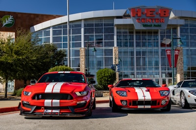 Two bright red sports cars with white racing stripes are parked in front of a large building with glass windows and a sign reading 'H-E-B Center at Cedar Park'. The sports cars contrast sharply against the clear blue sky and the modern architectural design of the building. There are trees and flagpoles in the background, adding a dynamic urban setting to the scene.