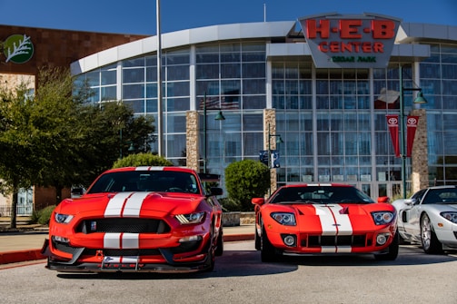 Two bright red sports cars with white racing stripes are parked in front of a large building with glass windows and a sign reading 'H-E-B Center at Cedar Park'. The sports cars contrast sharply against the clear blue sky and the modern architectural design of the building. There are trees and flagpoles in the background, adding a dynamic urban setting to the scene.