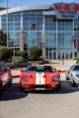 A red sports car with white racing stripes is parked in front of a modern building with large glass windows. The building has stone pillars and is labeled as the H-E-B Center. There are other vehicles parked beside the sports car and green shrubs are visible around the area.