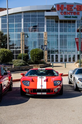 A red sports car with white racing stripes is parked in front of a modern building with large glass windows. The building has stone pillars and is labeled as the H-E-B Center. There are other vehicles parked beside the sports car and green shrubs are visible around the area.