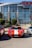A red sports car with white racing stripes is parked in front of a modern building with large glass windows. The building has stone pillars and is labeled as the H-E-B Center. There are other vehicles parked beside the sports car and green shrubs are visible around the area.