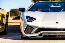 A sleek, modern sports car with a white exterior is prominently displayed. The car's design features sharp lines and aggressive styling typical of high-performance vehicles. In the background, there is a glimpse of another car with a yellow finish, suggesting a setting of luxury and automotive excellence.