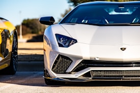 A sleek, modern sports car with a white exterior is prominently displayed. The car's design features sharp lines and aggressive styling typical of high-performance vehicles. In the background, there is a glimpse of another car with a yellow finish, suggesting a setting of luxury and automotive excellence.
