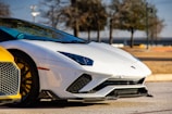 A sleek sports car with a white body, featuring angular design elements and distinctive headlights. The car is parked on a paved surface with trees in the background. A portion of a yellow car is visible in the foreground, showcasing a honeycomb grille and stylish alloy wheels.