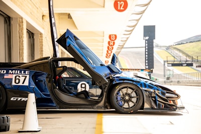 A sleek, blue race car is positioned in a pit stop area. The car's gullwing door is open, revealing a detailed and high-tech interior. The number 67 is prominently displayed on the side, alongside branded decals. The setting includes a clear view of the racetrack to the side, with lap timing displays and consecutive pit stop bays above, each numbered.