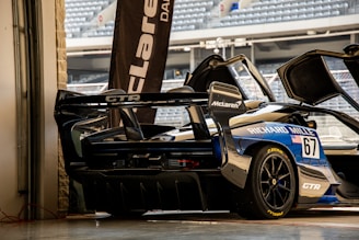 A high-performance sports car with a sleek design is parked in a garage setting. The vehicle features prominent sponsorship logos, including McLaren and Richard Mille. The garage door is open, revealing a glimpse of stadium seating in the background.