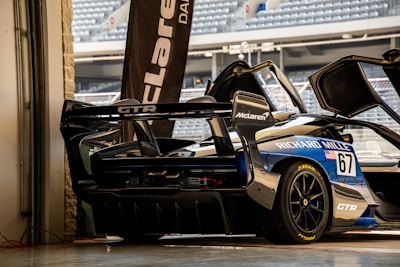 A high-performance sports car with a sleek design is parked in a garage setting. The vehicle features prominent sponsorship logos, including McLaren and Richard Mille. The garage door is open, revealing a glimpse of stadium seating in the background.