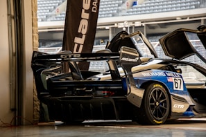 A high-performance sports car with a sleek design is parked in a garage setting. The vehicle features prominent sponsorship logos, including McLaren and Richard Mille. The garage door is open, revealing a glimpse of stadium seating in the background.