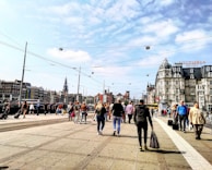 A busy urban street scene with a crowd of people walking on a wide pavement. Classic European architecture is visible in the background, including a large historic building with a sign for Victoria Hotel. Overhead, tram cables crisscross the sky under scattered clouds. The atmosphere is vibrant with people carrying bags and suitcases, suggesting a bustling travel or commute area.