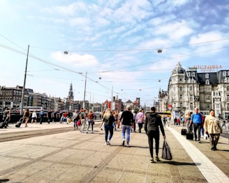 A busy urban street scene with a crowd of people walking on a wide pavement. Classic European architecture is visible in the background, including a large historic building with a sign for Victoria Hotel. Overhead, tram cables crisscross the sky under scattered clouds. The atmosphere is vibrant with people carrying bags and suitcases, suggesting a bustling travel or commute area.