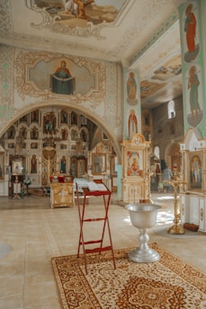 The image depicts the interior of an ornately decorated church. The walls and ceilings are adorned with religious paintings and icons. A red stand holds an open book, possibly a Bible, near a decorative rug. A large, ornate silver baptismal font is prominently visible. The overall atmosphere is serene and sacred, emphasized by the intricate designs and warm lighting.
