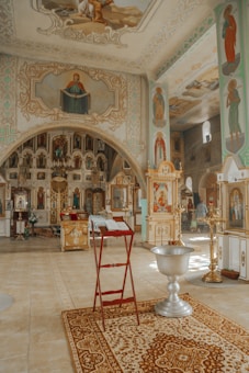 The image depicts the interior of an ornately decorated church. The walls and ceilings are adorned with religious paintings and icons. A red stand holds an open book, possibly a Bible, near a decorative rug. A large, ornate silver baptismal font is prominently visible. The overall atmosphere is serene and sacred, emphasized by the intricate designs and warm lighting.