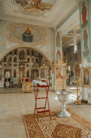 The image depicts the interior of an ornately decorated church. The walls and ceilings are adorned with religious paintings and icons. A red stand holds an open book, possibly a Bible, near a decorative rug. A large, ornate silver baptismal font is prominently visible. The overall atmosphere is serene and sacred, emphasized by the intricate designs and warm lighting.