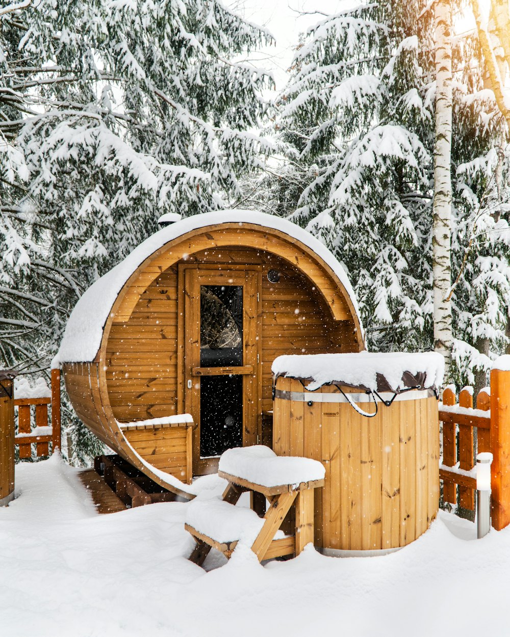 Wood-fired barrel sauna and cold plunge tub covered in snow, with snow falling