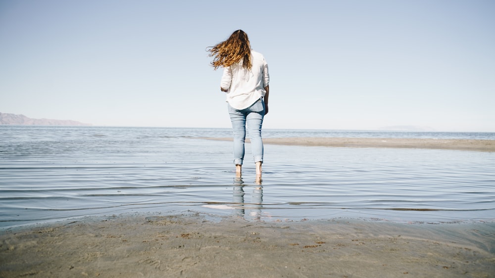 woman in white long sleeve shirt and blue denim jeans standing on beach during daytime